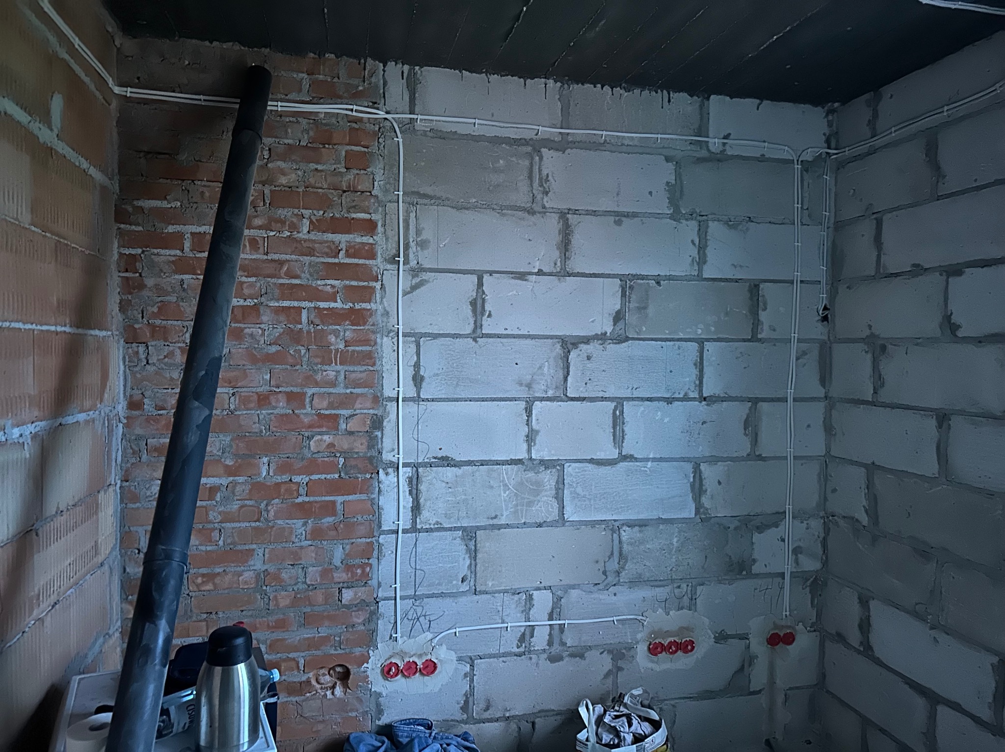 Wewnętrzna instalacja elektryczna w surowym stanie, widoczne białe przewody prowadzone po ścianie z betonu komórkowego i cegły, czerwone puszki pod gniazdka, czarna rura wentylacyjna przy ścianie...