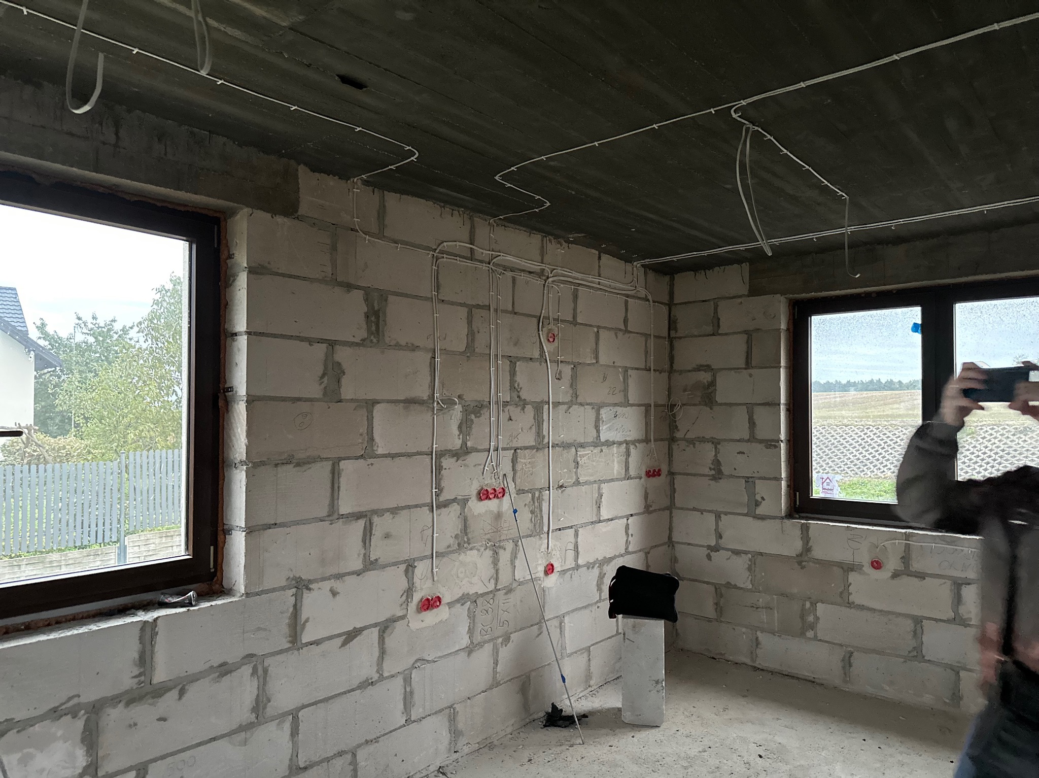 Surowy stan wnętrza z widoczną instalacją elektryczną: przewody poprowadzone po ścianach z bloczków betonowych, puszki elektryczne w kolorze czerwonym, okna z brązowymi ramami, betonowy sufit...