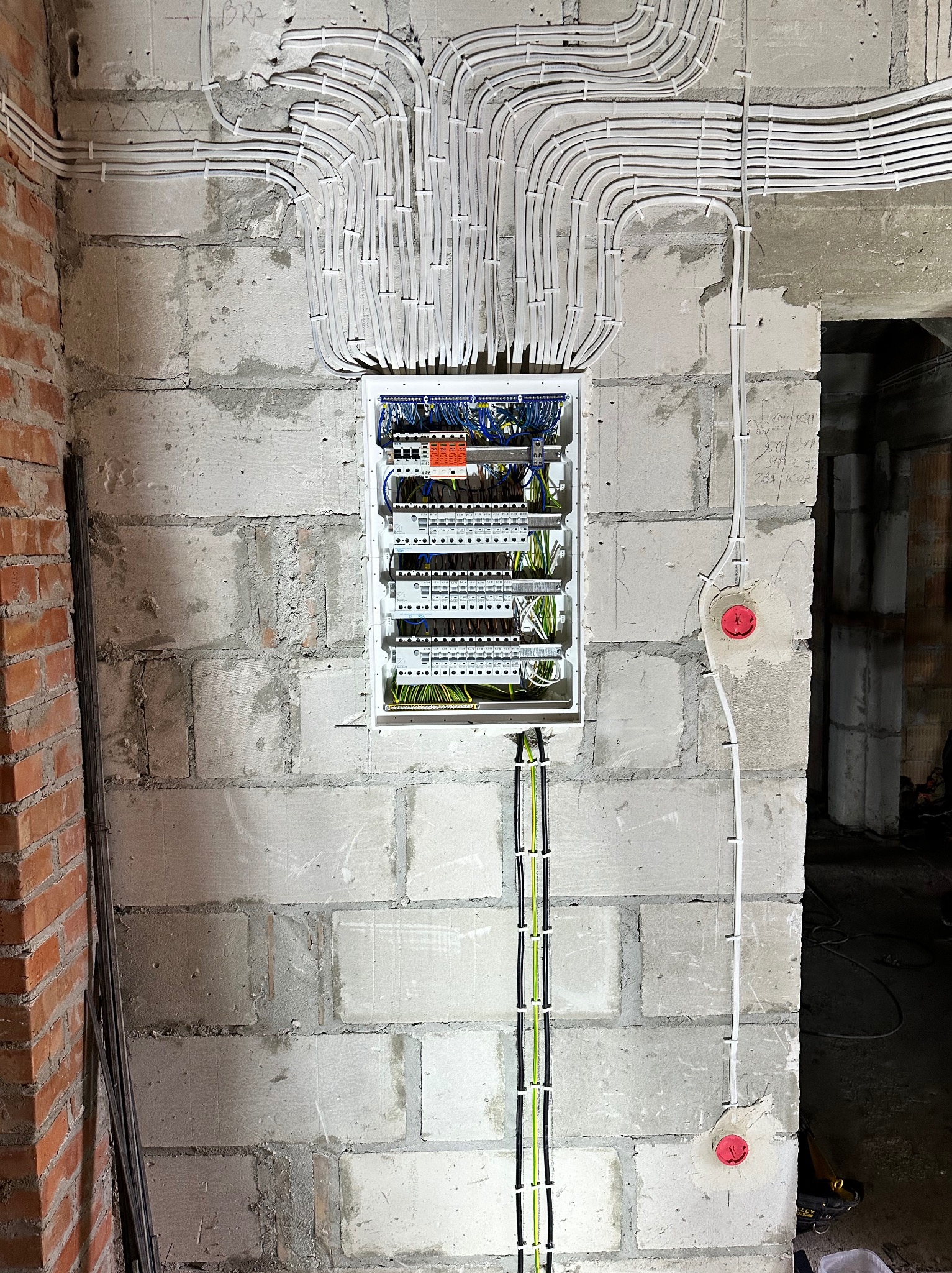 Rozdzielnica elektryczna z licznymi przewodami ułożonymi w peszlach i przymocowanymi do surowej ściany z betonu komórkowego, widoczne także gniazda elektryczne z czerwonymi zaślepkami.