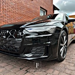 Fura na błysk - Błyszczący, czarny Audi A6 po detailingu, z widoczną kratką grilla i reflektorem, na tle ceglanej ściany i kostki brukowej. Efekt hydrofobowy na lakierze.