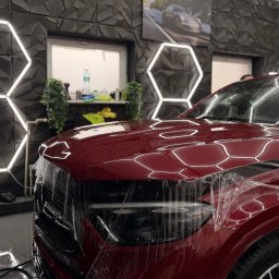Fura na błysk - Folie PPF & Detailing Łódź - Czerwony samochód w trakcie zabezpieczania folią ochronną PPF w profesjonalnym studiu detailingu. Widoczne oświetlenie heksagonalne i dekoracyjna ściana.