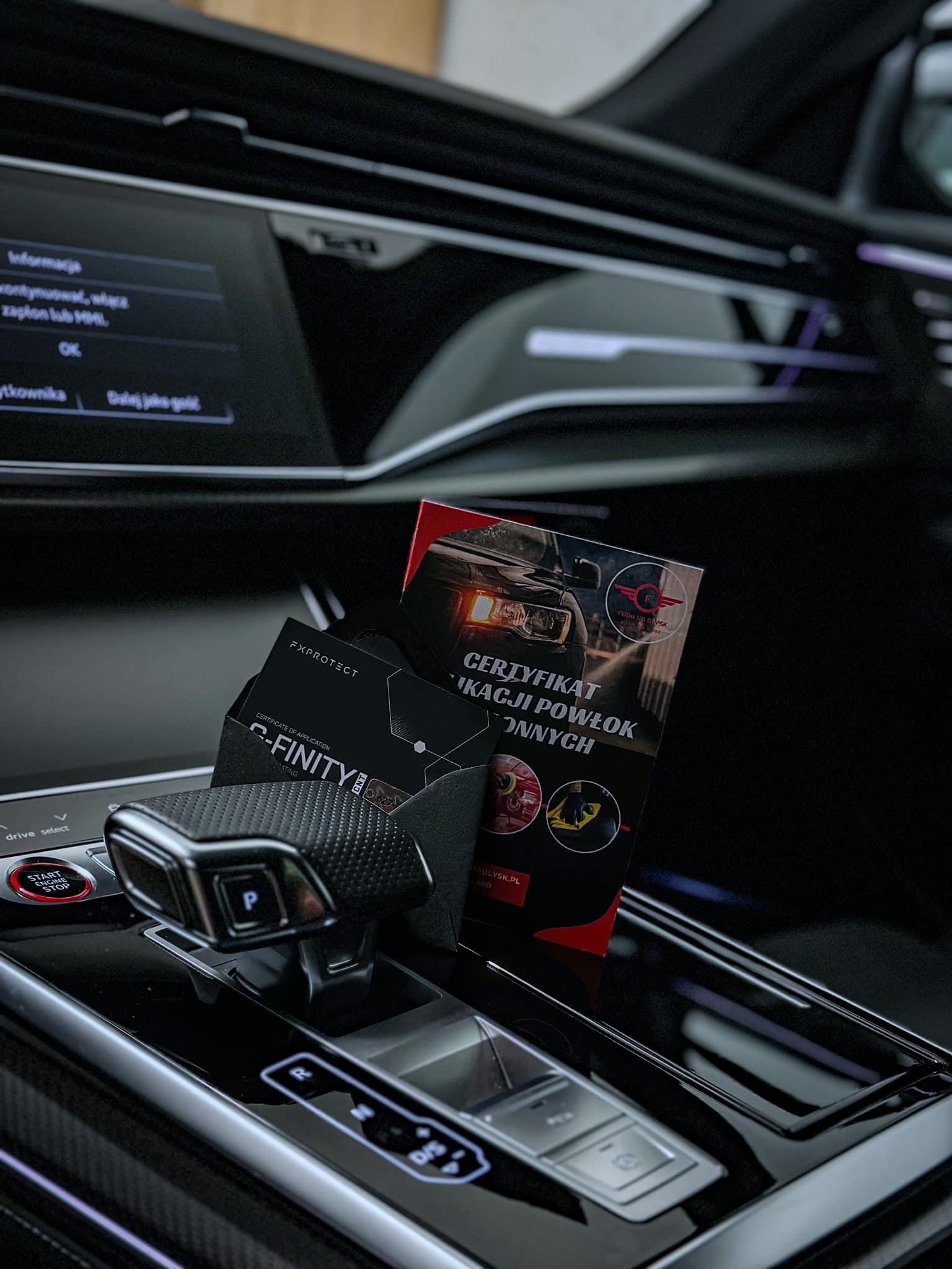 Wnętrze samochodu z czarną deską rozdzielczą, na której leży certyfikat aplikacji powłok ochronnych marki FX Protect oraz wizytówka firmy detailingowej.