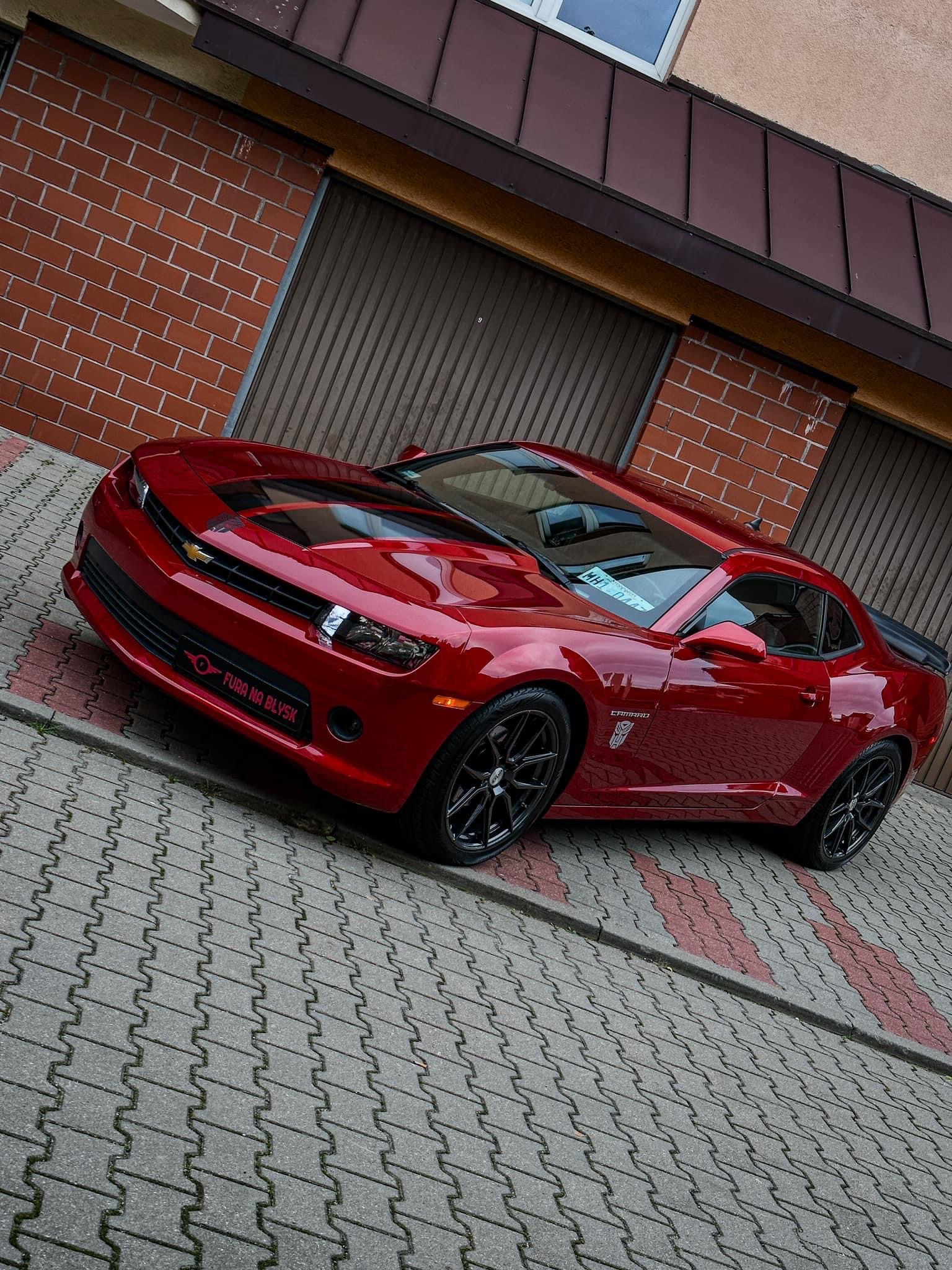 Czerwony Chevrolet Camaro po detailingu, z czarnymi pasami na masce i naklejką 'Fura na Błysk' na przednim zderzaku, zaparkowany przed garażem z ceglaną ścianą.