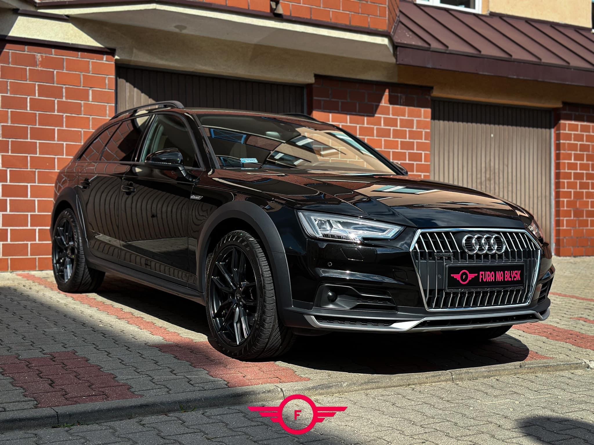 Czarny samochód Audi Allroad po myciu, z widocznym logo 'Fura na Błysk' na tablicy rejestracyjnej, zaparkowany na kostce brukowej przed budynkiem z czerwonej cegły.