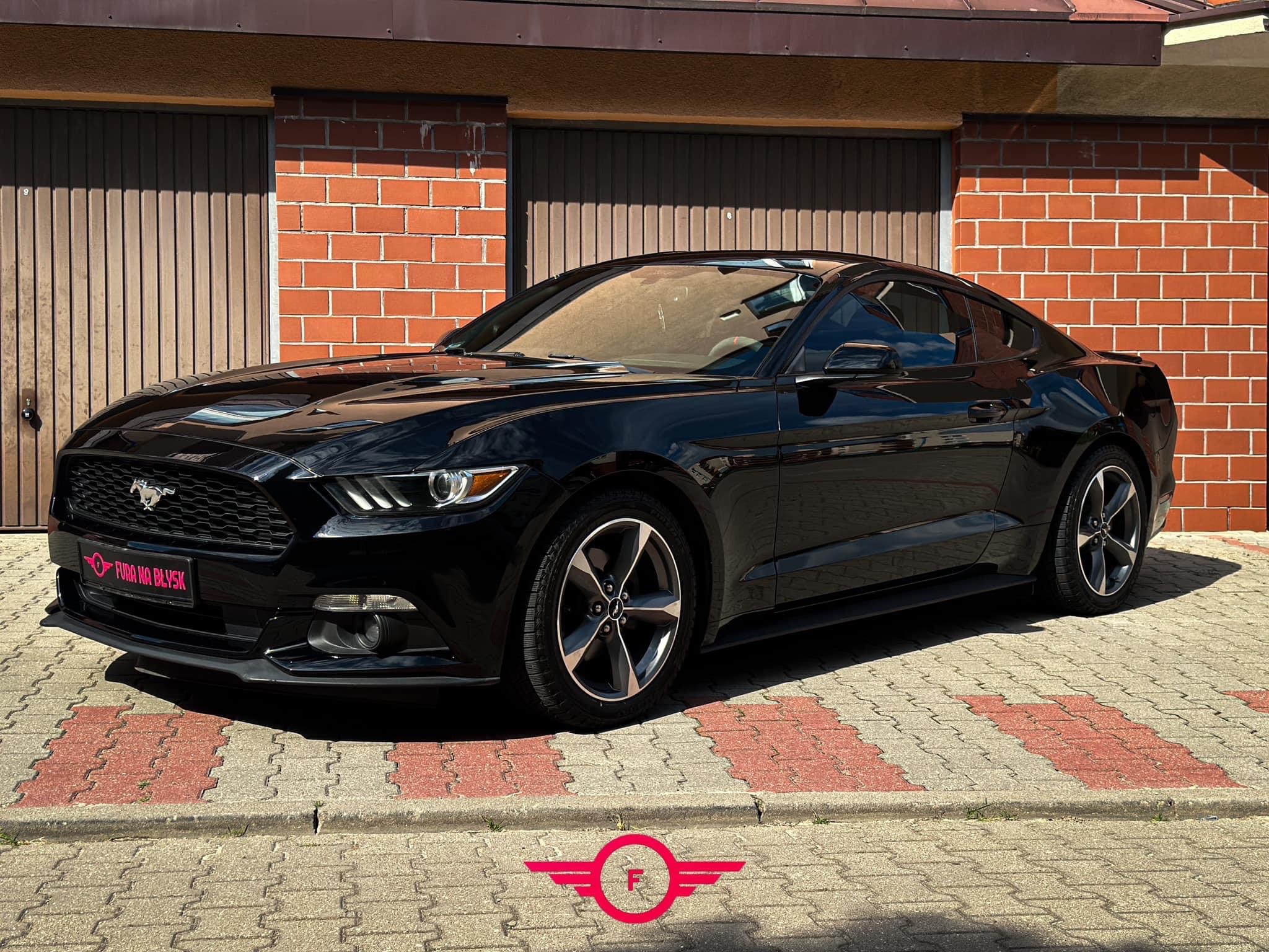 Błyszczący, czarny Ford Mustang zaparkowany na kostce brukowej przed garażem z ceglaną ścianą, widoczny napis 'Fura na błysk' na przednim zderzaku.