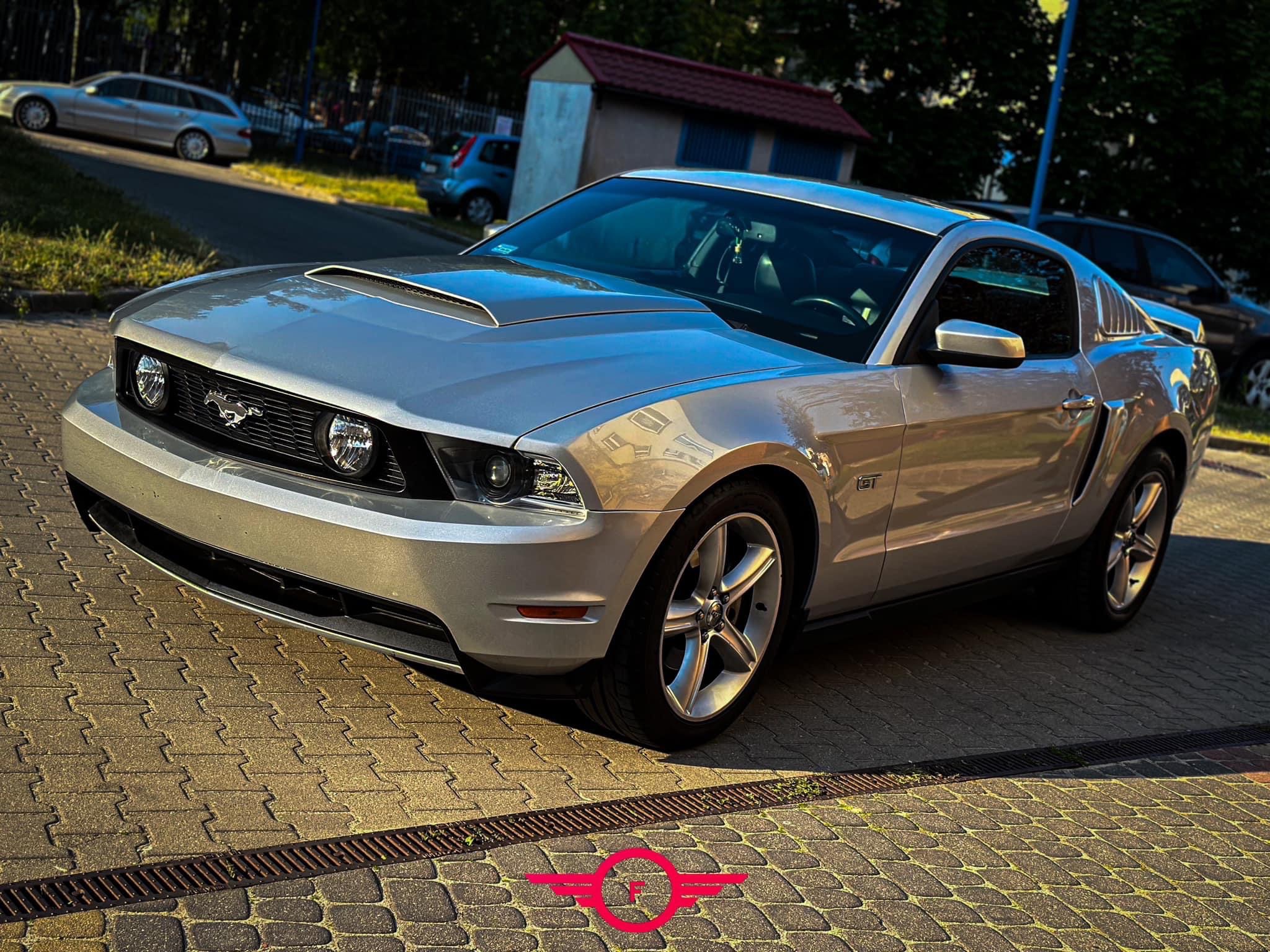 Srebrny Ford Mustang GT zaparkowany na brukowanym podjeździe, widoczny z przodu pod kątem, z budynkiem w tle.