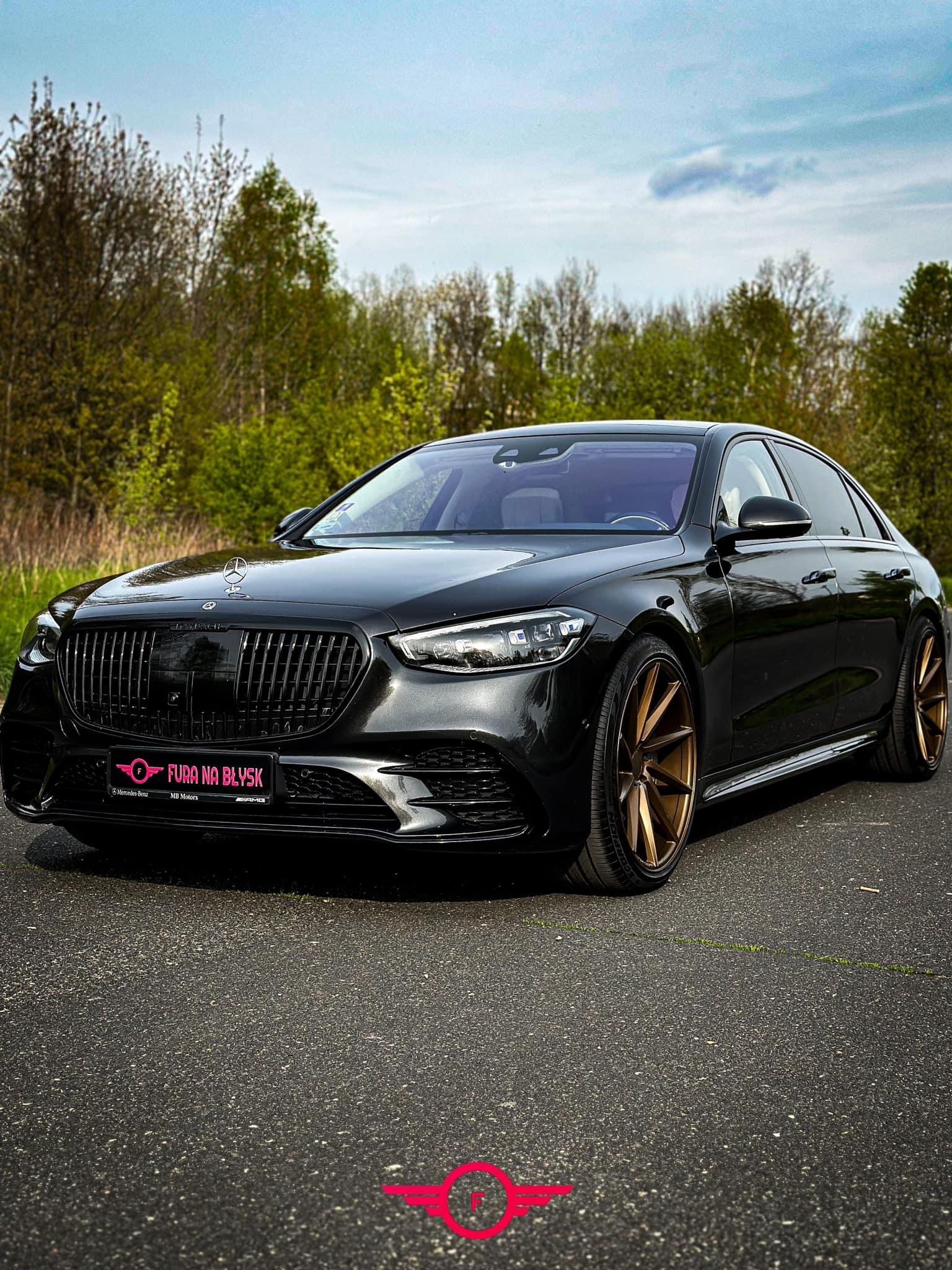 Czarna limuzyna Mercedes-Benz S-Class po tuningu z ciemnymi felgami w kolorze brązu, na tle drzew i nieba.