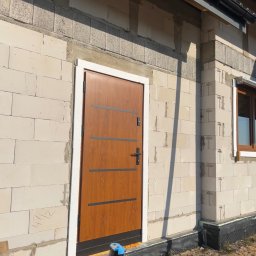 Vekline Agnieszka Markowska - Brązowe drzwi zewnętrzne z poziomymi, ciemnymi paskami, zamontowane w surowym murze z białych bloczków, widoczne fragmenty okna i orynnowania, na dole zabezpieczenie przeciwwilgociowe i fragment...