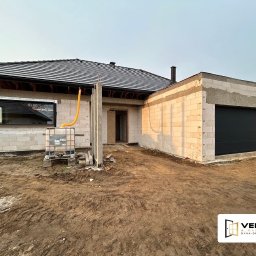 Vekline Agnieszka Markowska - Nowoczesny dom w trakcie budowy z czarną bramą garażową, oknem PCV i elementami konstrukcyjnymi, ukazujący stan surowy zamknięty z widocznym zbiornikiem na wodę.