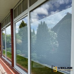 Vekline Agnieszka Markowska - Białe okna PCV z żaluzjami plisowanymi, widok z zewnątrz. Czerwona cegła na elewacji. Widoczne odbicie nieba i zieleni w szybach.