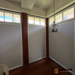 Vekline Agnieszka Markowska - Zasłonięte okna plisami w jasnym wnętrzu z ceglanym filarem. Widoczna lampa wisząca i fragment plecionego krzesła. Ciemna podłoga.