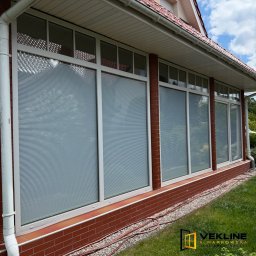 Vekline Agnieszka Markowska - Białe okna z żaluzjami wewnętrznymi w ceglanej elewacji domu. Widoczne przewody na trawie i logo firmy w rogu kadru. Ujęcie z dołu, słoneczny dzień.