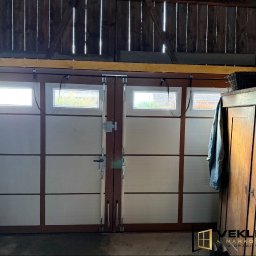 Vekline Agnieszka Markowska - Biała brama garażowa segmentowa z oknami, widok z wnętrza garażu. Drewniana konstrukcja stropu, szafa w tle, logo firmy w prawym dolnym rogu.