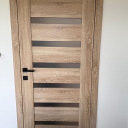 Vekline Agnieszka Markowska - Drewniane drzwi wewnętrzne z poziomymi przeszkleniami i czarną klamką, widoczne na tle białej ściany. Wykończenie w odcieniach dębu.