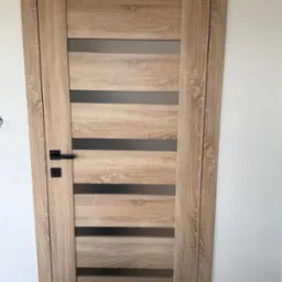 Drewniane drzwi wewnętrzne z poziomymi przeszkleniami i czarną klamką, widoczne na tle białej ściany. Wykończenie w odcieniach dębu.