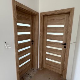 Vekline Agnieszka Markowska - Dwa zamontowane, drewniane drzwi wewnętrzne z przeszkleniami w poziomych pasach, czarne klamki, widoczne kable wystające ze ściany.