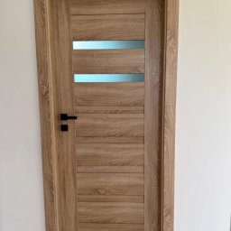 Vekline Agnieszka Markowska - Drewniane drzwi wewnętrzne z poziomymi przeszkleniami i czarną klamką, zamontowane w białej ścianie. Widoczny fragment podłogi i kabel zasilający.