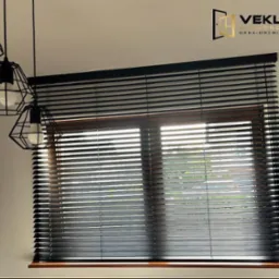 Ciemne, drewniane żaluzje poziome zamontowane w oknie z drewnianą ramą. W tle widoczne światło dzienne. U góry zwisają trzy czarne lampy w stylu loft.