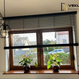 Vekline Agnieszka Markowska - Drewniane okno z czarnymi żaluzjami poziomymi, przez które widać domy i zieleń. Na parapecie dwa kwiaty w doniczkach. Nad oknem logo firmy VEKLINE.