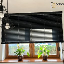 Vekline Agnieszka Markowska - Czarne żaluzje poziome zamontowane w oknie z drewnianą ramą. Na parapecie stoją dwie doniczki z roślinami. Nad oknem wiszą lampy w geometrycznych kloszach.