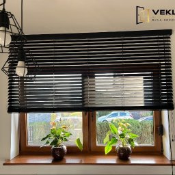 Vekline Agnieszka Markowska - Ciemne żaluzje poziome w oknie z drewnianą ramą, z dwoma kwiatami w doniczkach na parapecie. Widoczny fragment lampy i logo firmy.