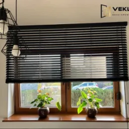 Ciemne żaluzje poziome w oknie z drewnianą ramą, z dwoma kwiatami w doniczkach na parapecie. Widoczny fragment lampy i logo firmy.