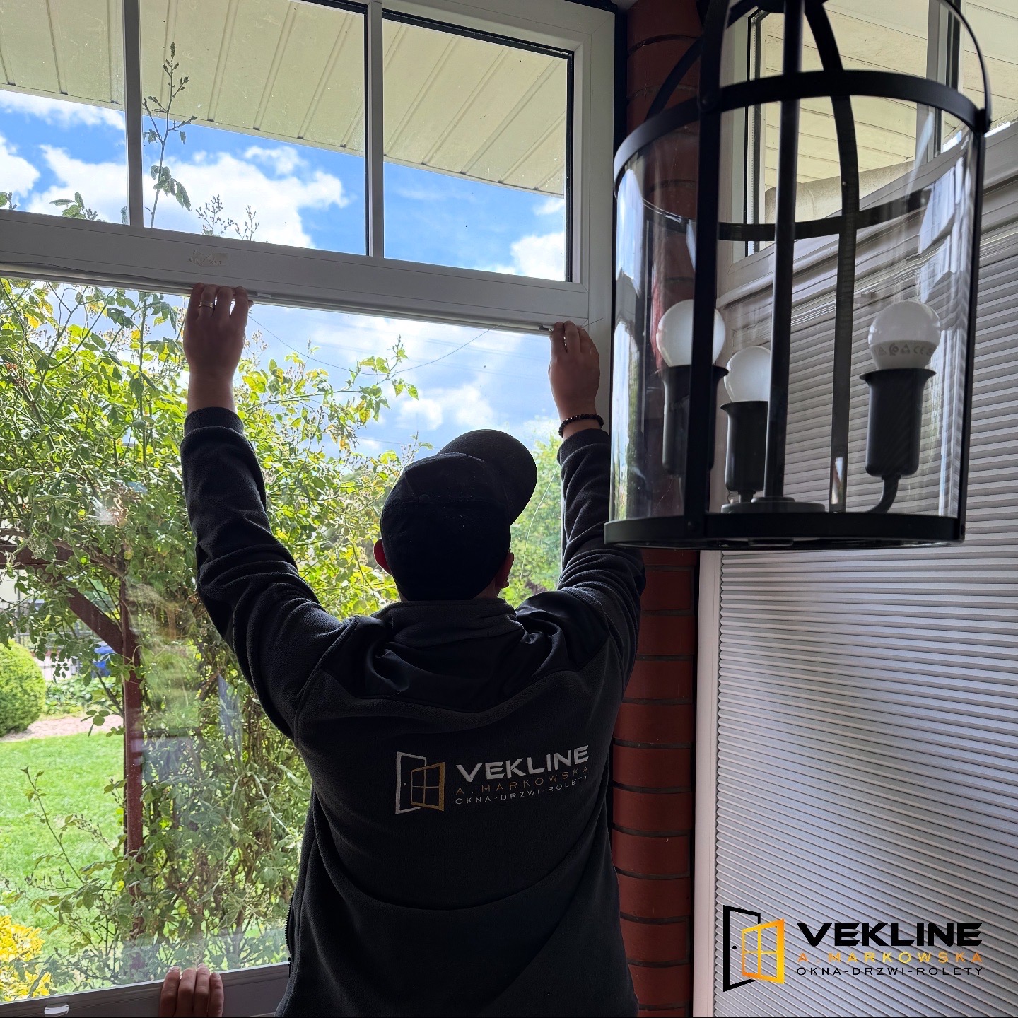 Mężczyzna w bluzie z logo Vekline montuje białe okno. Widok zza pleców, na pierwszym planie lampa. Za oknem zieleń i błękitne niebo.
