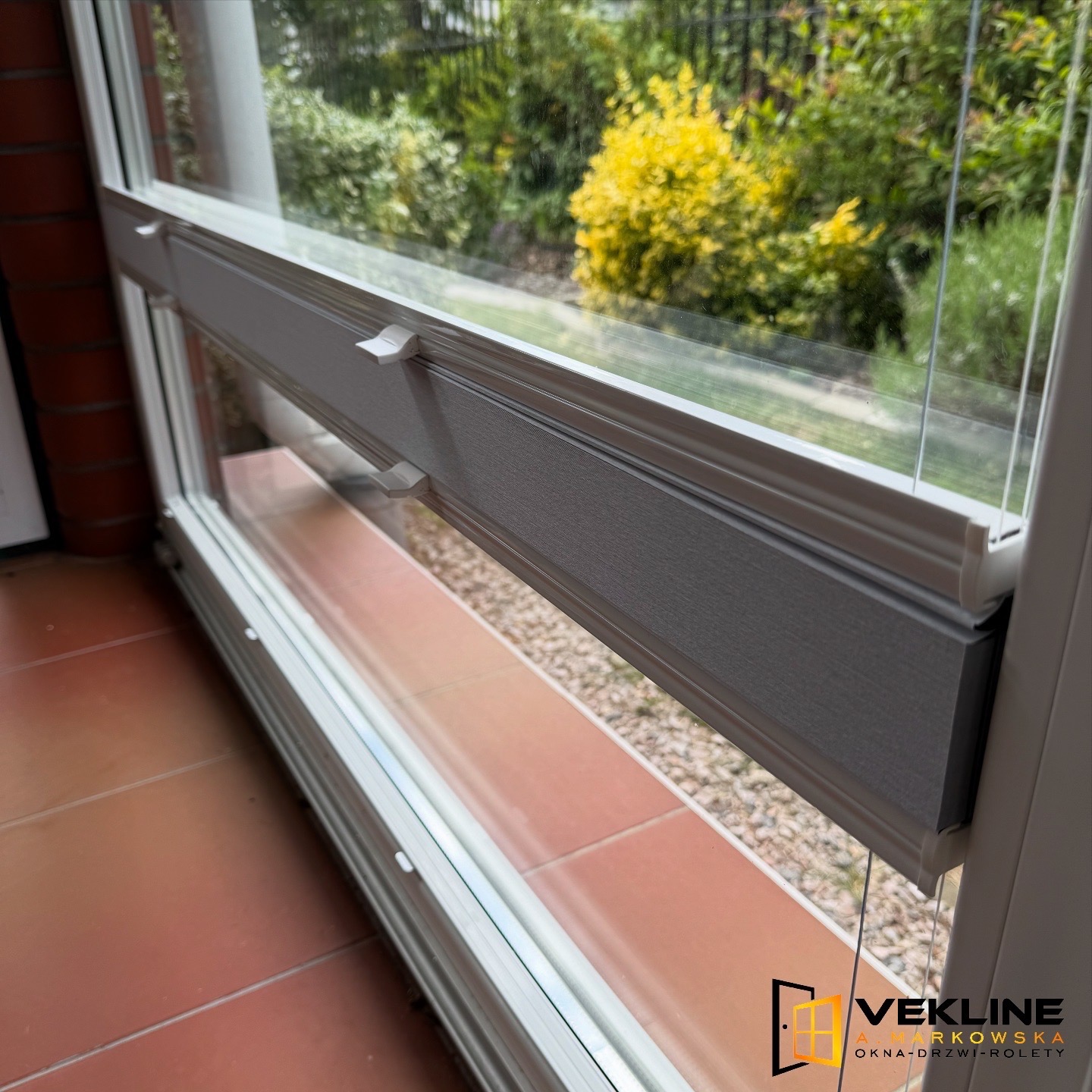 Szare rolety plisowane zamontowane w oknie PCV, widok z bliska na mechanizm i teksturę materiału, z zielenią ogrodu w tle.