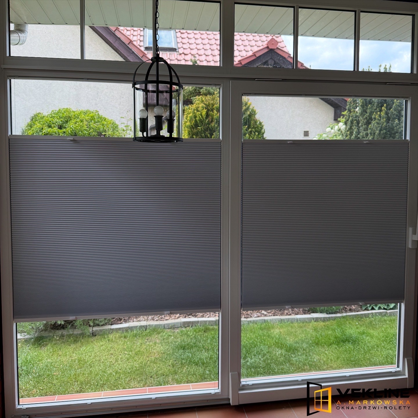 Szare, plisowane rolety w oknach z widokiem na ogród, z dekoracyjną lampą wiszącą. Widoczne detale okien i faktura rolet.