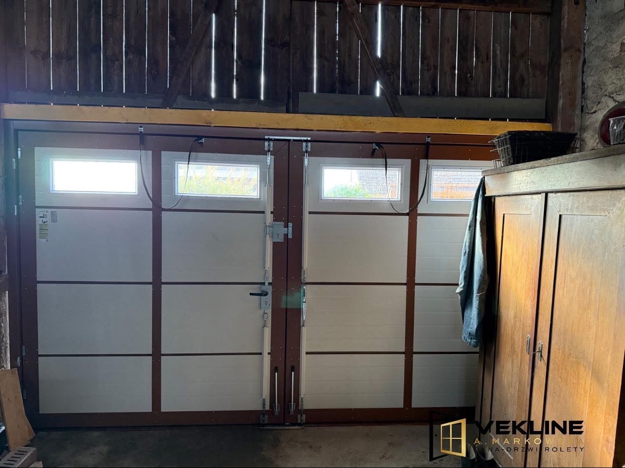 Biała brama garażowa segmentowa z oknami, widok z wnętrza garażu. Drewniana konstrukcja stropu, szafa w tle, logo firmy w prawym dolnym rogu.