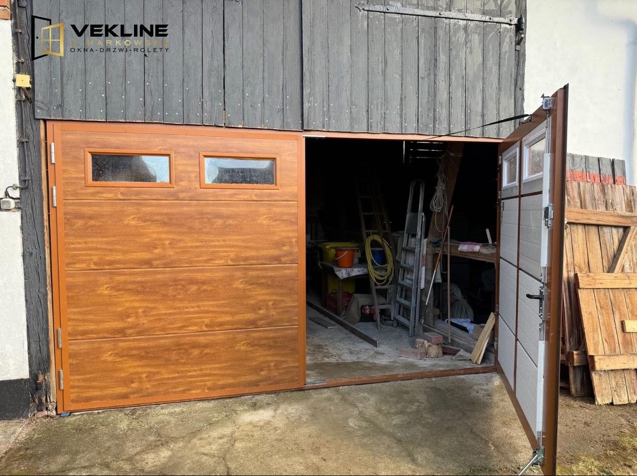 Otwarta garażowa brama segmentowa w kolorze drewna, z dwoma małymi okienkami, zamontowana w budynku o szarej, drewnianej elewacji. Widoczne wnętrze garażu.