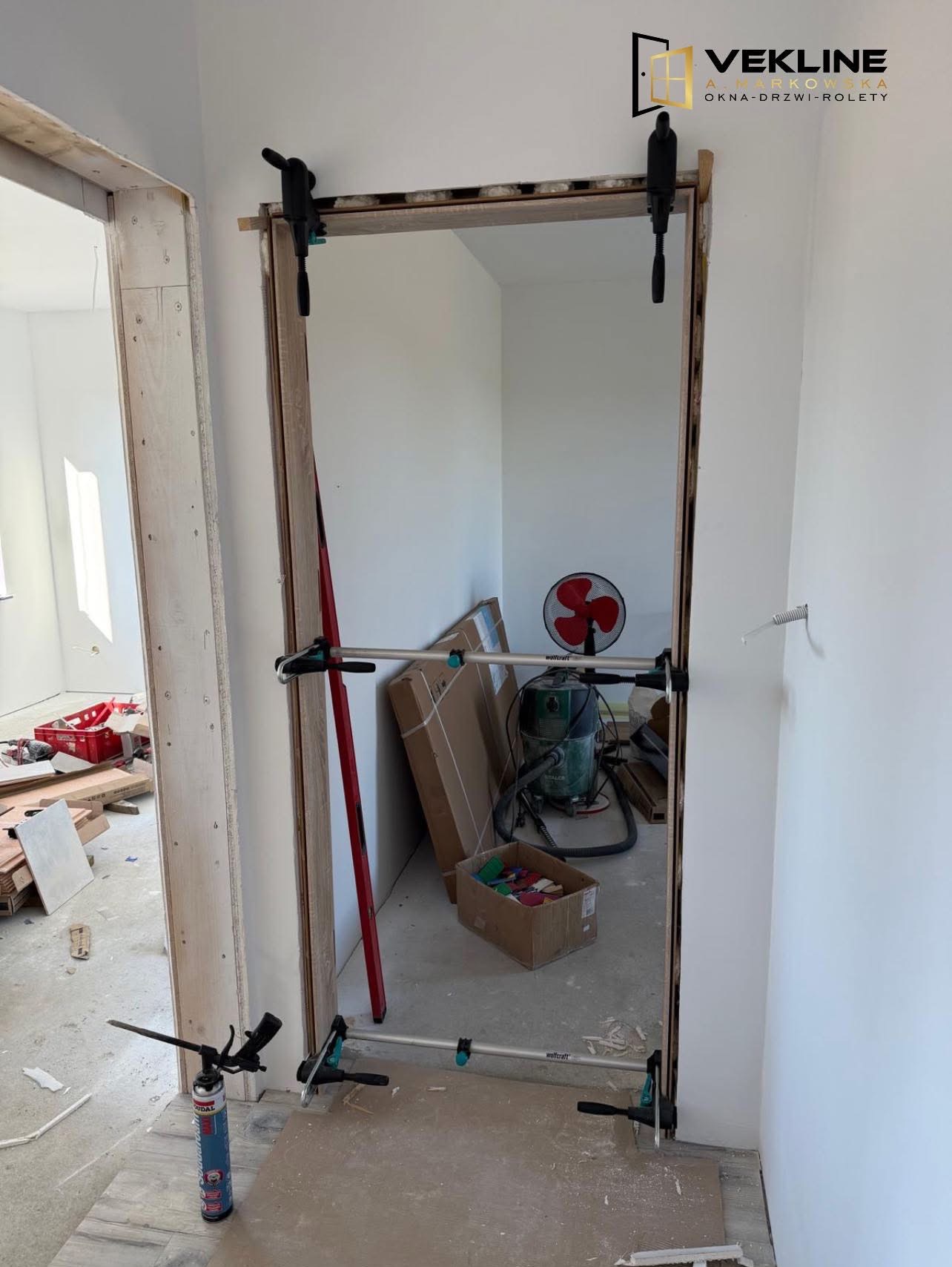 Montaż ościeżnicy drzwiowej z użyciem rozpór i klamer, widoczne narzędzia i materiały budowlane w surowym wnętrzu. Częściowo widoczny wentylator.