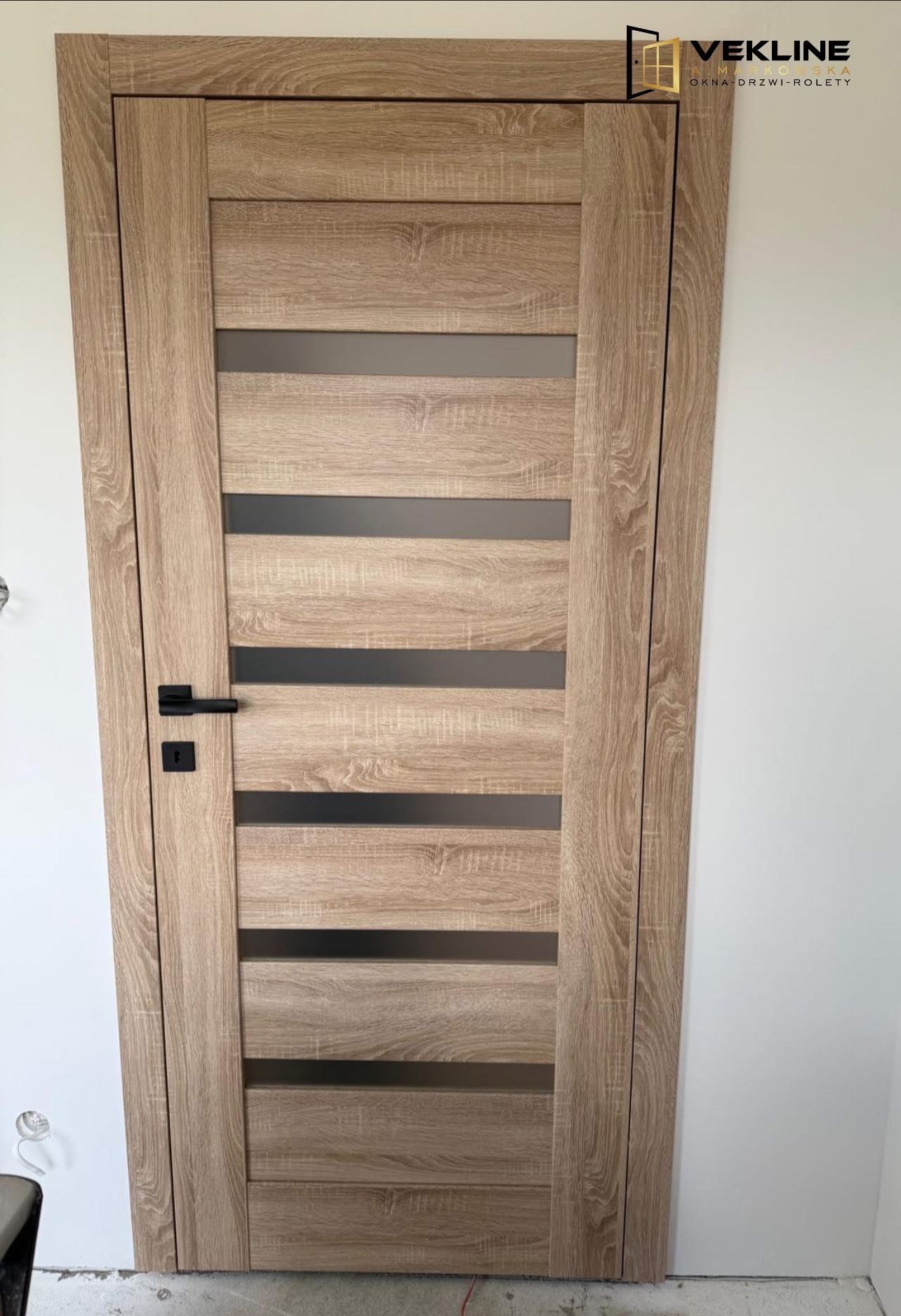 Drewniane drzwi wewnętrzne z poziomymi przeszkleniami i czarną klamką, widoczne na tle białej ściany. Wykończenie w odcieniach dębu.