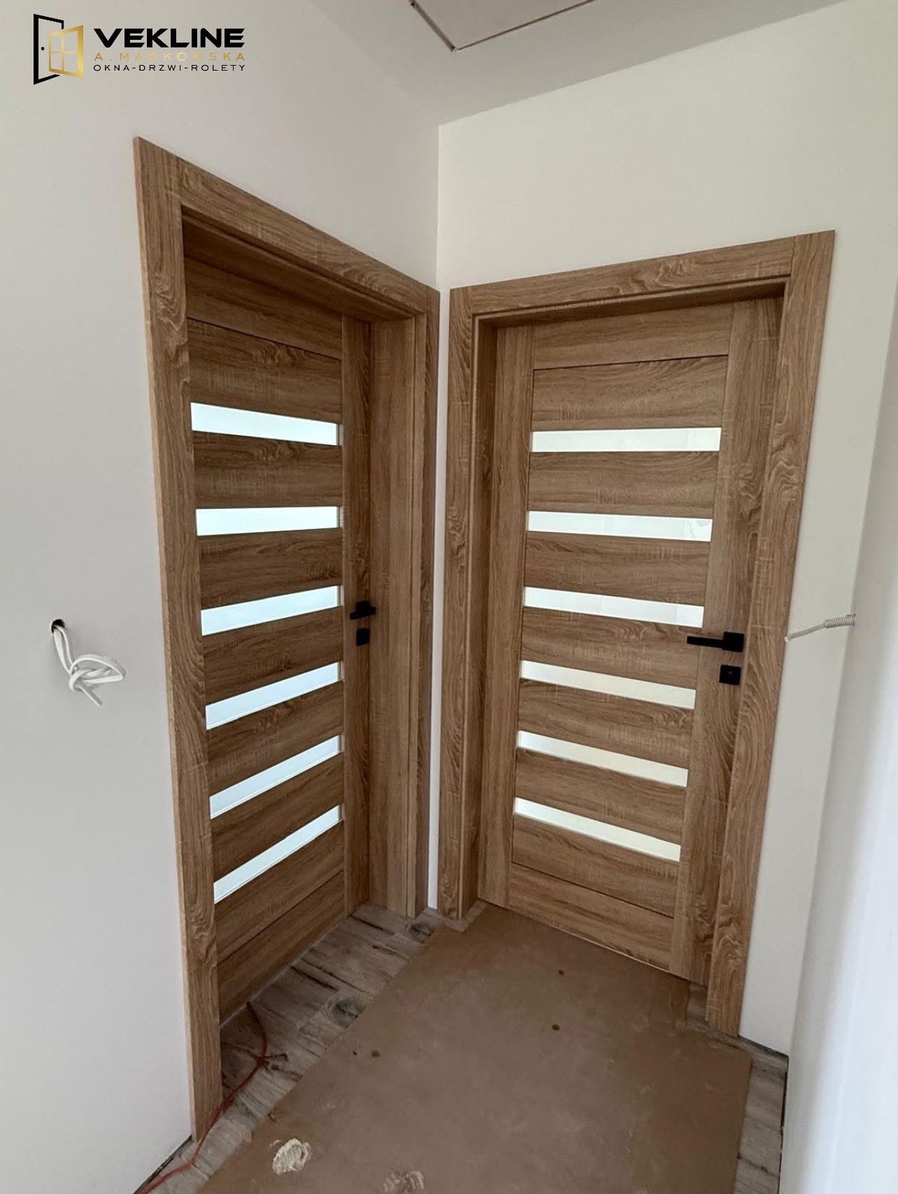 Dwa zamontowane, drewniane drzwi wewnętrzne z przeszkleniami w poziomych pasach, czarne klamki, widoczne kable wystające ze ściany.