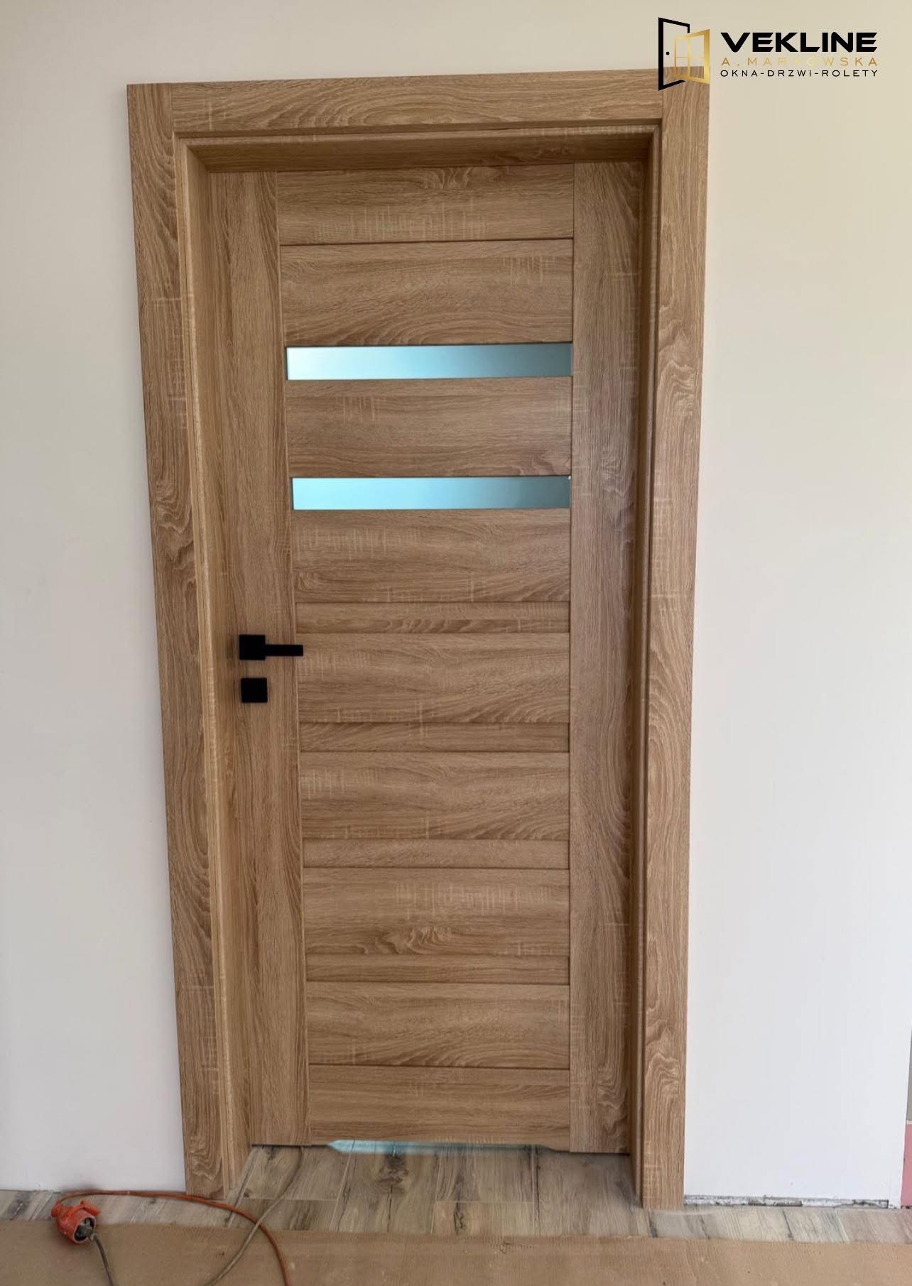 Drewniane drzwi wewnętrzne z poziomymi przeszkleniami i czarną klamką, zamontowane w białej ścianie. Widoczny fragment podłogi i kabel zasilający.
