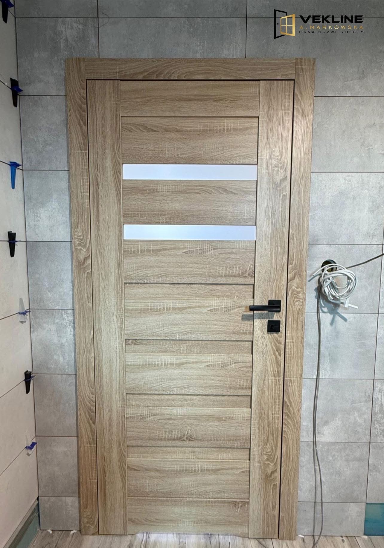 Beżowe drzwi wewnętrzne z poziomymi przeszkleniami, czarny klamka, szare płytki na ścianie, widoczne narzędzia montażowe i logo firmy Vekline w prawym górnym rogu.