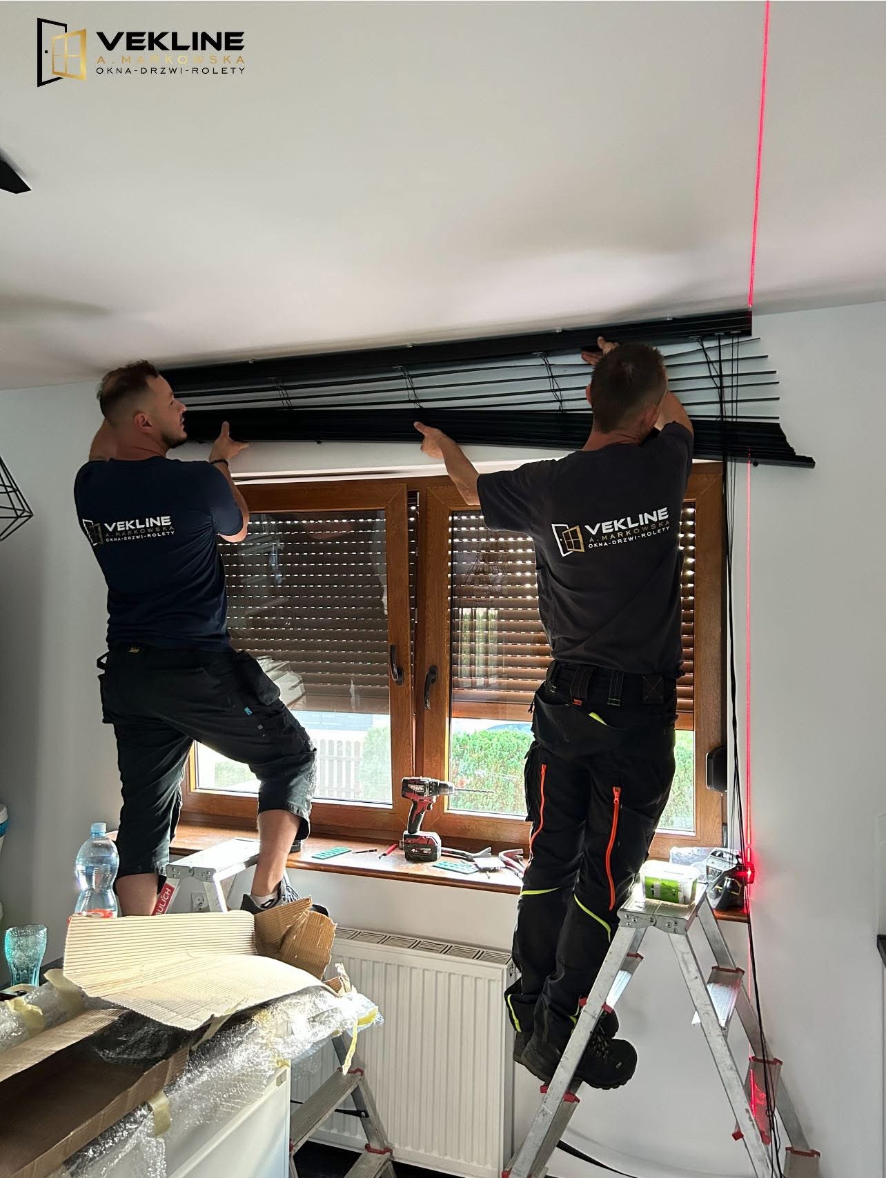 Dwóch monterów na drabinach instaluje czarne żaluzje poziome przy oknie z brązową ramą, używając poziomicy laserowej. Widoczne narzędzia i logo firmy na koszulkach.