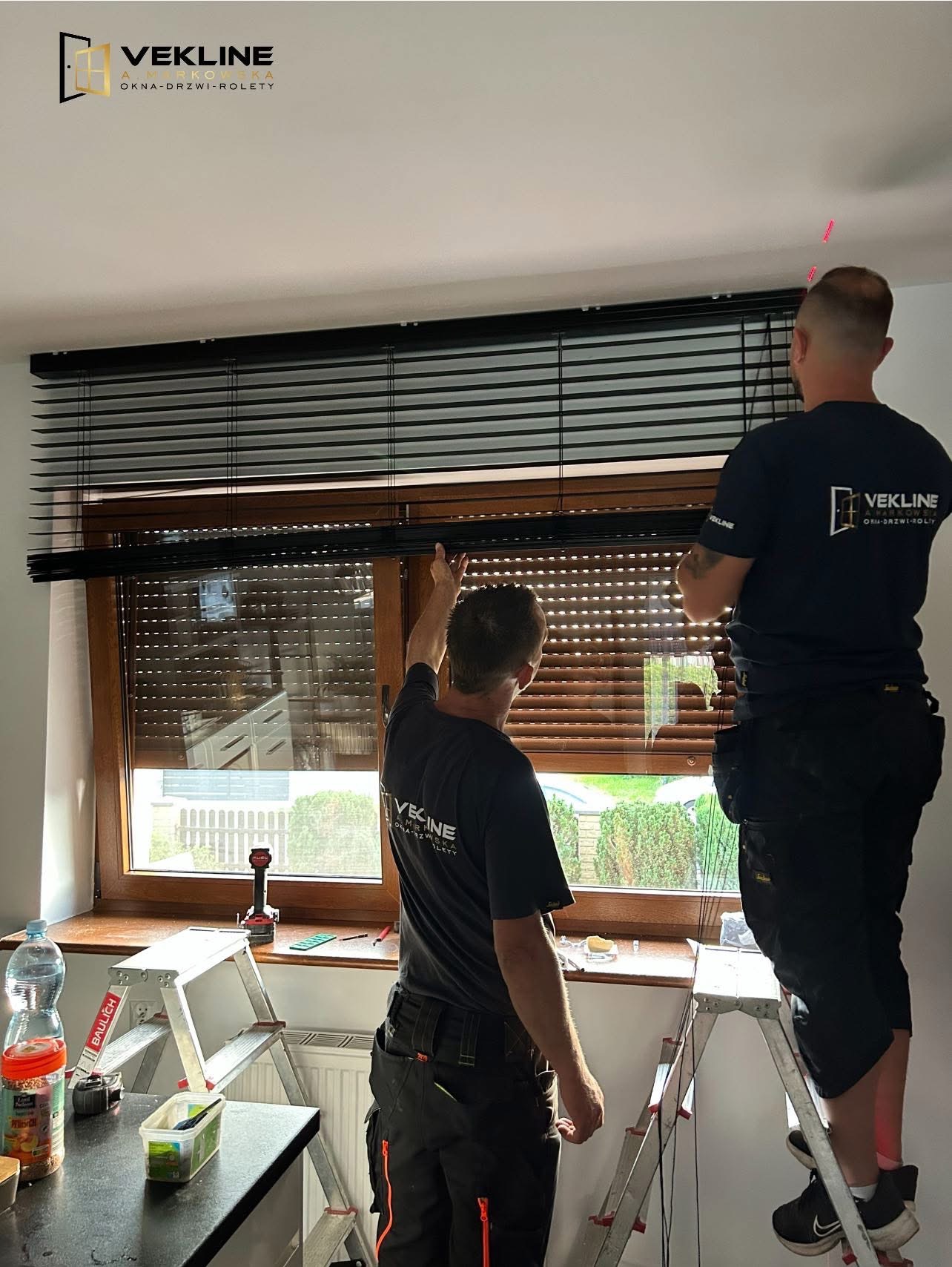 Montaż żaluzji poziomych przez dwóch monterów w firmowych koszulkach. Widoczne narzędzia i drabina. Okno z roletą zewnętrzną w tle.