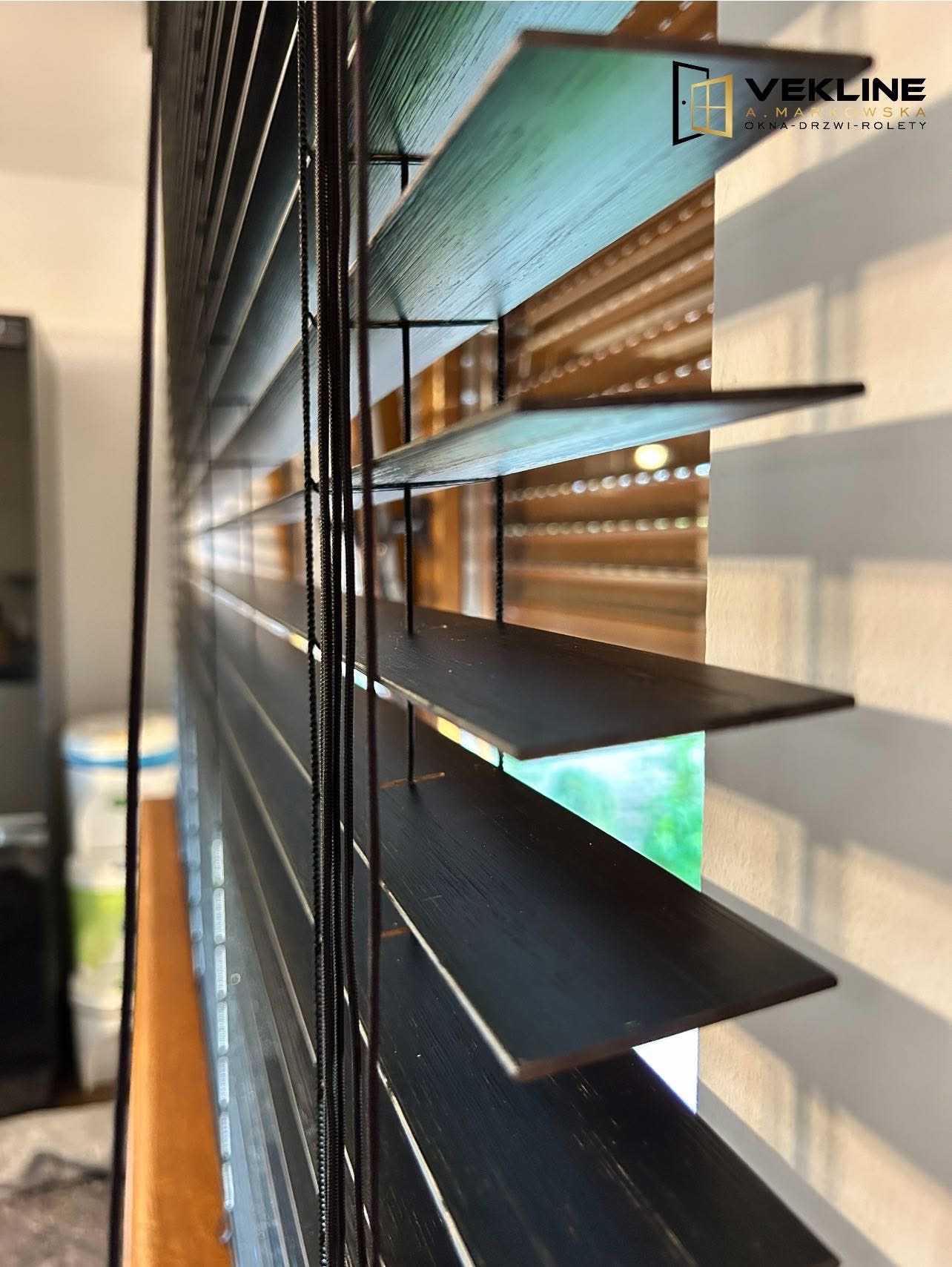 Ciemne, drewniane żaluzje poziome, widok z boku, ukazujące detale konstrukcji i regulację światła. Logo firmy w prawym górnym rogu.