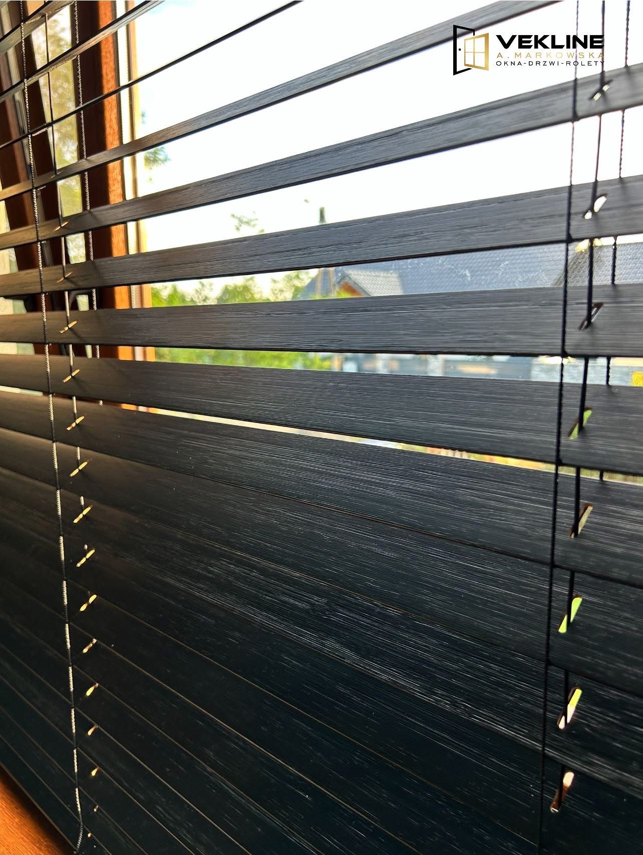 Ciemne, drewniane żaluzje poziome z widocznym logotypem firmy w prawym górnym rogu. Delikatne światło dzienne prześwituje przez szpary.
