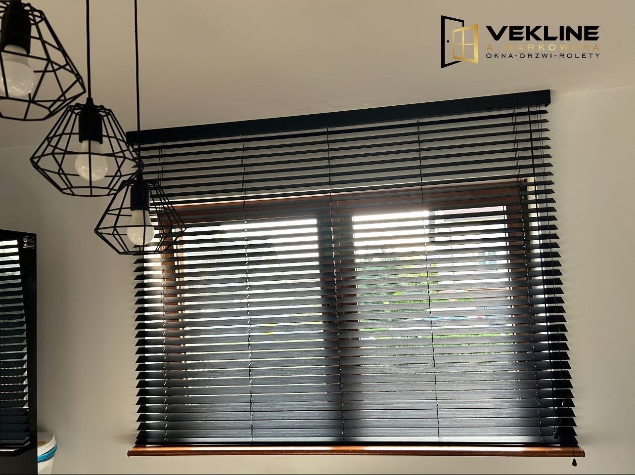 Ciemne, drewniane żaluzje poziome zamontowane w oknie z drewnianą ramą. W tle widoczne światło dzienne. U góry zwisają trzy czarne lampy w stylu loft.