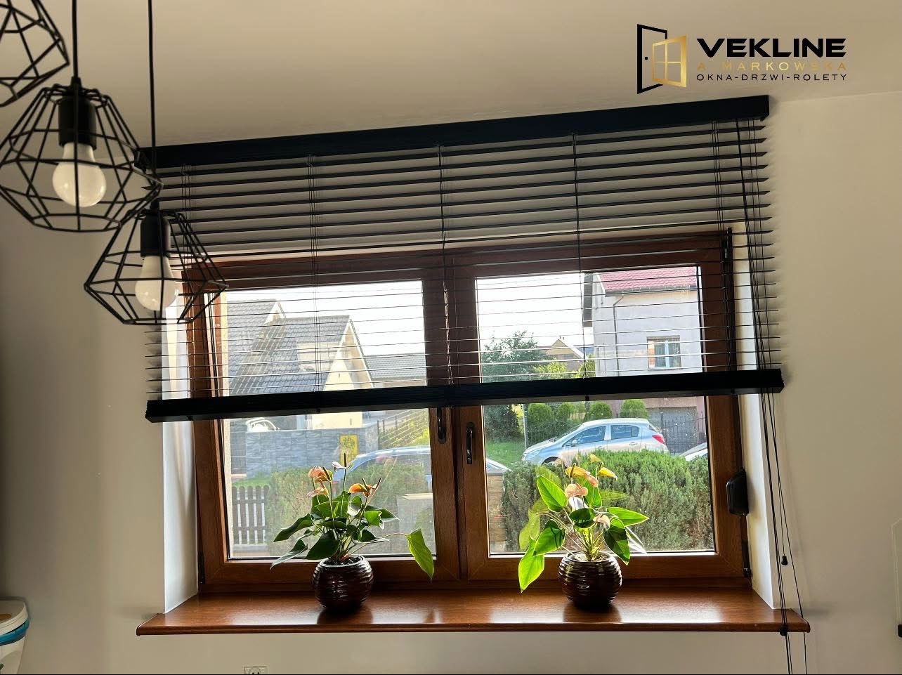 Drewniane okno z czarnymi żaluzjami poziomymi, przez które widać domy i zieleń. Na parapecie dwa kwiaty w doniczkach. Nad oknem logo firmy VEKLINE.