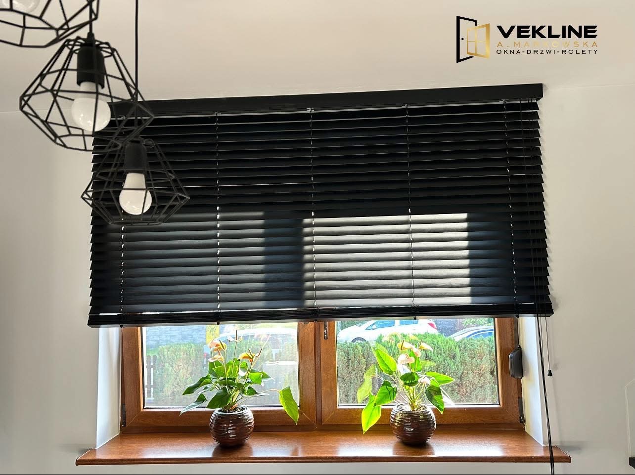 Czarne żaluzje poziome zamontowane w oknie z drewnianą ramą. Na parapecie stoją dwie doniczki z roślinami. Nad oknem wiszą lampy w geometrycznych kloszach.