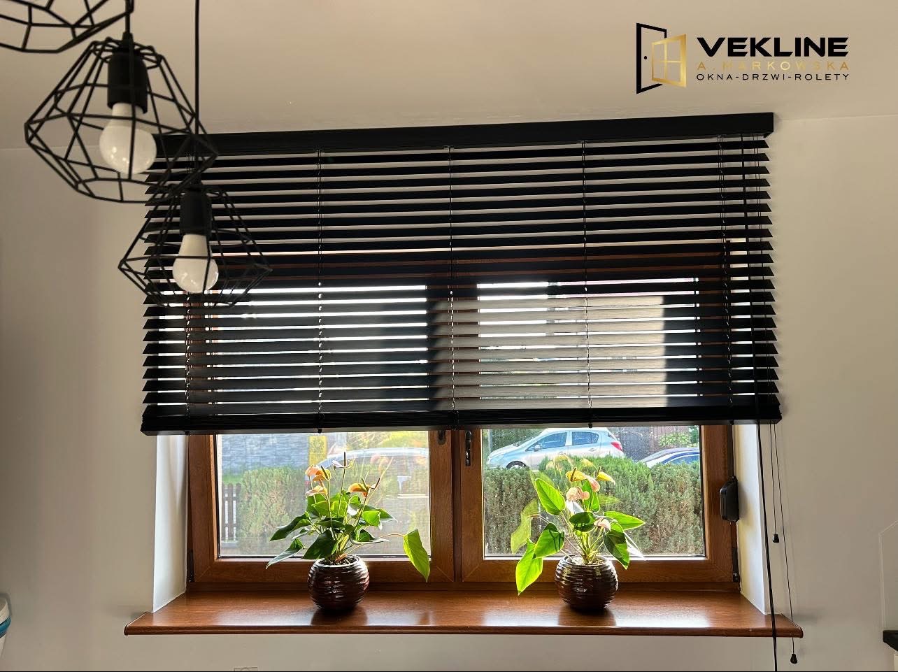 Ciemne żaluzje poziome w oknie z drewnianą ramą, z dwoma kwiatami w doniczkach na parapecie. Widoczny fragment lampy i logo firmy.