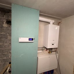 W-MAX Waldemar Wróblewski - Biały piec gazowy wiszący na ścianie w surowym pomieszczeniu, widoczne rury, skrzynka z instalacją wodną, oraz płyta gipsowo-kartonowa z zamontowanym sterownikiem z wyświetlaczem.