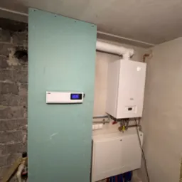 Biały piec gazowy wiszący na ścianie w surowym pomieszczeniu, widoczne rury, skrzynka z instalacją wodną, oraz płyta gipsowo-kartonowa z zamontowanym sterownikiem z wyświetlaczem.