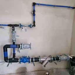 W-MAX Waldemar Wróblewski - Złożona instalacja wodna z rur niebieskich i czarnych, z licznymi zaworami i filtrami, zamontowana na surowej, szarej ścianie w pomieszczeniu technicznym.