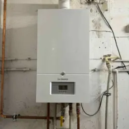 Biały, wiszący piec gazowy De Dietrich na tle surowej, pomalowanej ściany z widocznymi rurami miedzianymi i plastikowymi, wentylator wyciągowy w górnym rogu, żółty zawór gazowy.