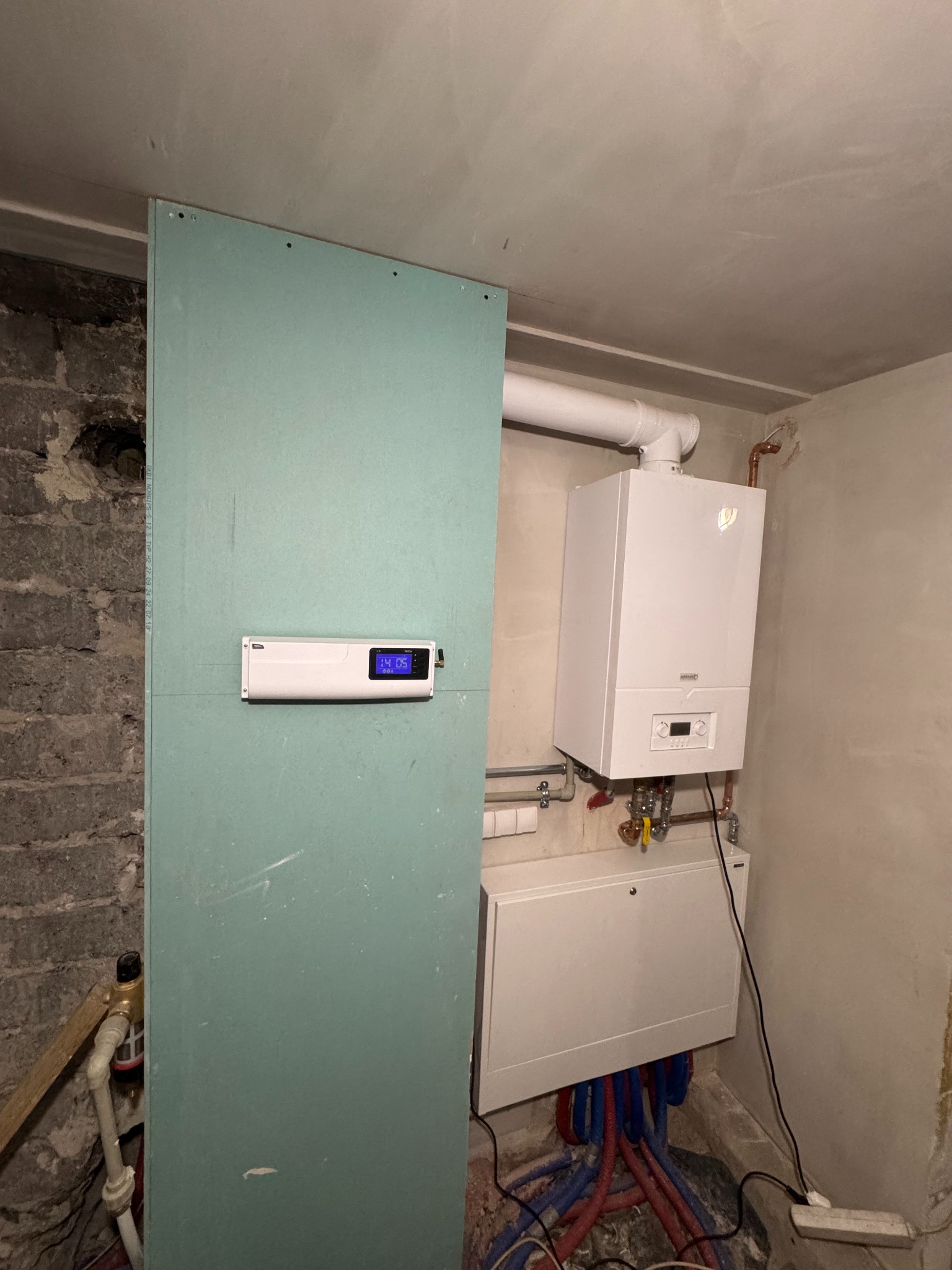 Biały piec gazowy wiszący na ścianie w surowym pomieszczeniu, widoczne rury, skrzynka z instalacją wodną, oraz płyta gipsowo-kartonowa z zamontowanym sterownikiem z wyświetlaczem.