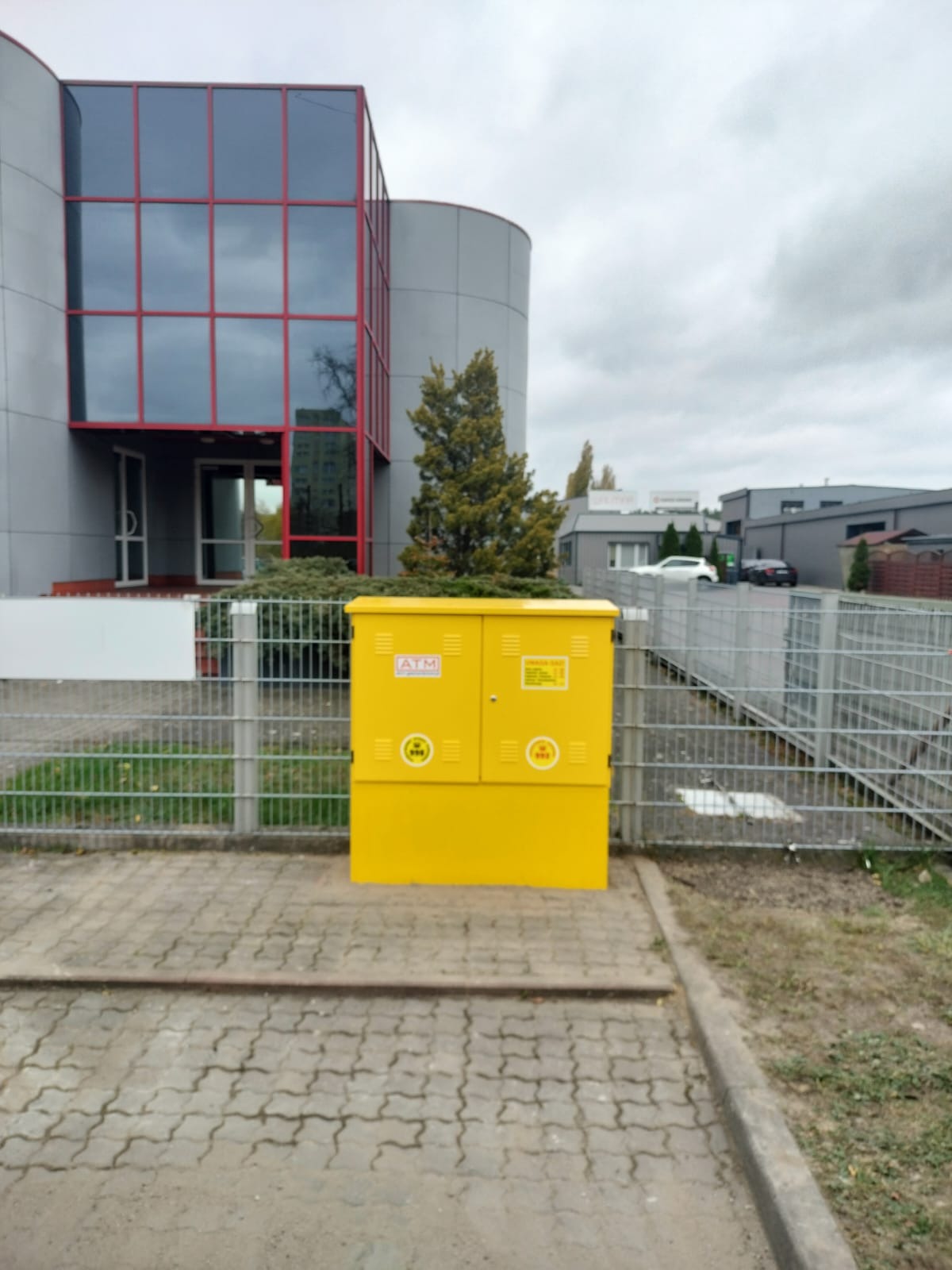 Jasnożółta szafka gazowa ATM na kostce brukowej, przed nowoczesnym budynkiem biurowym z czerwonymi ramami okiennymi, oddzielona metalowym ogrodzeniem.